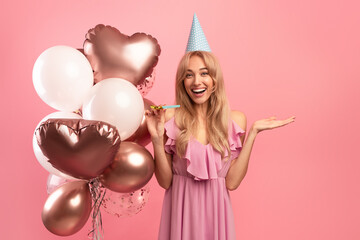 Obraz na płótnie Canvas Young blonde lady with festive balloons, birthday cap and party blower celebrating special occasion on pink background