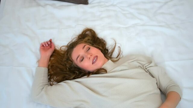 Traveling, living in hotels concept. Portrait of tired happy young woman with closed eyes is lying on bed in hotel after hard road, camera in motion. She is smiling and resting on white linen.