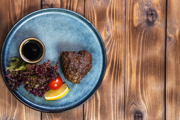 Grilled meat in a plate with lettuce, lemon and tomatoes close-up on a plate on a brown wooden background. Flat layout. Copy the space.