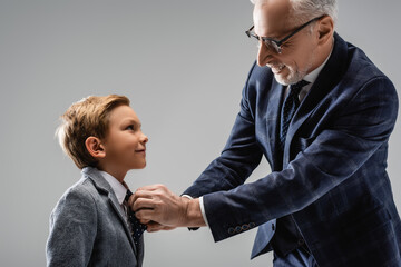 happy businessman in eyeglasses buttoning blazer of smiling grandson isolated on grey