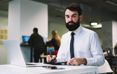 Serious bearded male manager writing notes in planner and looking at camera