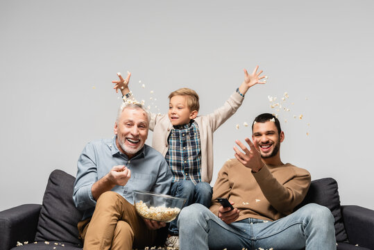 excited boy with dad and grandfather throwing popcorn while watching tv isolated on grey