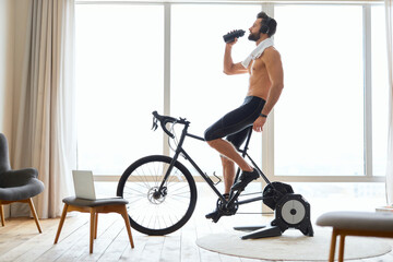Fototapeta na wymiar Athletic young man riding stationery bike and drinking water