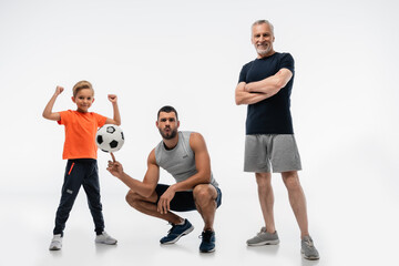 man in sportswear whistling while playing with soccer ball near boy and grandfather on white