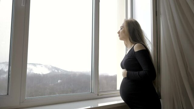 Young pregnant woman standing by the window and holding her belly