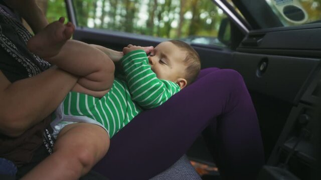 A little baby is lying in his mothers lap in a car as she is talking on the phone