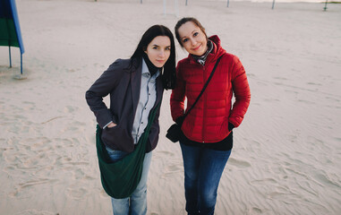 Obraz na płótnie Canvas Two young girls in blue jeans and jackets stand side by side and look into the frame at the viewer against the backdrop of coastal sand, beach.Concept of love between sisters, friendship and good mood