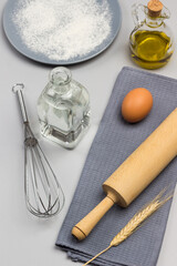 Rolling pin, egg and spikelet of wheat on gray napkin. Metal whisk, Two bottles of water and oil. Gray plate with flour