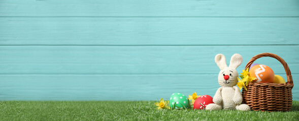 Colorful Easter eggs, basket and bunny toy on green grass against light blue wooden background, space for text. Horizontal banner design