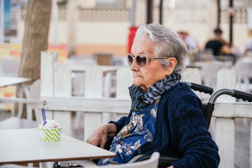 Elderly caucasian woman with pink sunglasses sitting on a wheelchair in a bar and with an ice cream