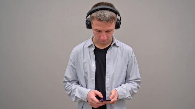 A young man on a gray background, in headphones, is typing text on a smartphone. Looks closely.