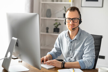 A portrait of young male wearing formal shirt using a headset and contemporary PC in the office, an...