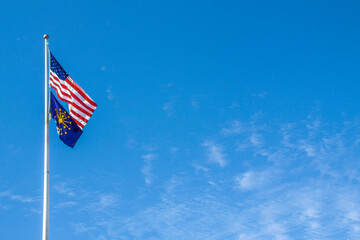 American and Indiana flags flying from tall flagpole against blue sky with whispy clouds - Room for copy