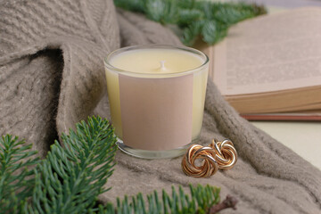 Eco-friendly handmade soy wax candle in a glass candlestick. Nearby lies a decor of fir branches, a warm sweater, a book and earrings.