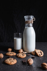 Glass and bottle with tasty milk placed with chocolate chip cookies and walnuts on table on black background for healthy breakfast 