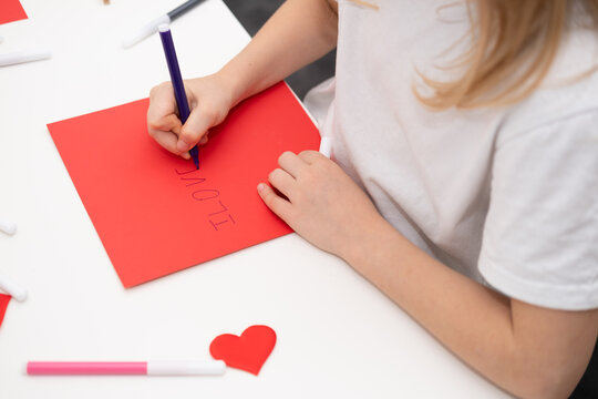 The child draws a postcard for the holiday of Mother's Day. Girl writes on red paper I love you mom
