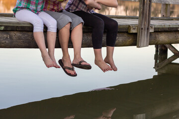 Three girls feet hanging from dock above water.