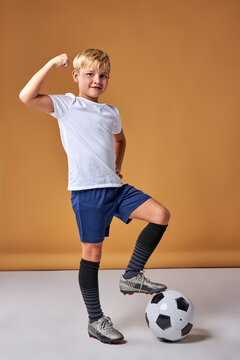 soccer player boy posing with ball in studio, blond kid boy stand un white and blue iniform isolated over brown background
