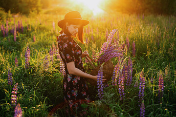 Fototapeta premium Beautiful woman relaxing in lupine field with rustic basket and flowers at sunset,tranquil moment