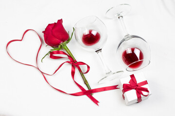 Fototapeta na wymiar Red heart of ribbon with red rose, wine glasses with red wine and gif box on white bed honeymoon. Surprise Valentine and wedding day in bed. Top view. Copy space.