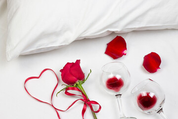 Red heart of ribbon with red rose, rose petals and wine glasses with red wine on white bed honeymoon. Surprise Valentine and wedding day in bed. Top view. Copy space