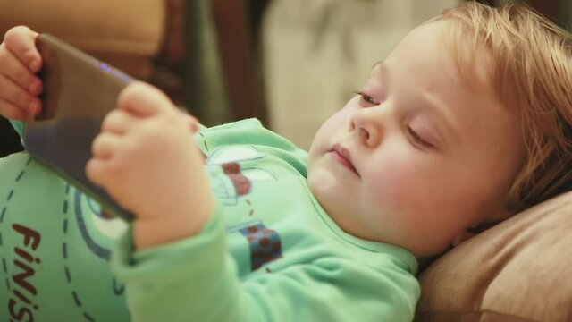 Charming baby boy with a new smartphone watching cartoons, lying on the couch at home. Close-up of a small kid with a phone resting in the bedroom. Selective focus