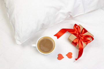 Obraz na płótnie Canvas Cup of coffee, gift box with red ribbon and little red heart on white bed with pillow. Morning romantic surprise flat lay background.