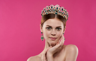 A romantic princess with a diadem on her head touches her face with her hands on a pink background