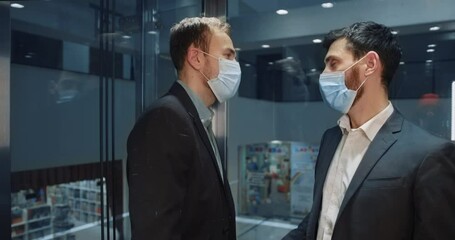 Two caucasian young corporate men in formal suits and facial masks moving to office inside glass elevator. Business people. Partnership. Lockdown.