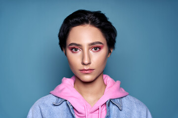 Confident stylish cool gen z teen girl, short-haired zoomer teenager with trendy pink makeup on pretty face wearing denim looking at camera isolated on blue background. Close up headshot portrait. - obrazy, fototapety, plakaty