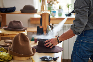 designer male designed created handmade hat, he steams hat with a steamer. in studio, at desktop....