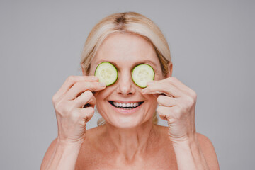 Cheerful healthy mature woman holding cucumber slices on eyes isolated over grey background