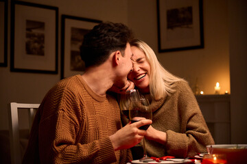 Happy young couple in love hugging, laughing, drinking wine, enjoying talking, having fun together celebrating Valentines day dining at home, having romantic dinner date with candles sitting at table. - Powered by Adobe