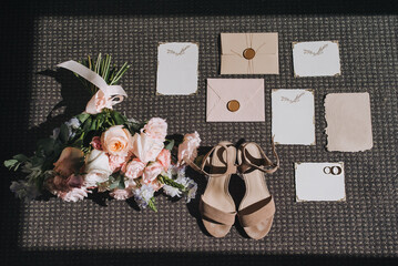 A large and beautiful bouquet of multi-colored roses, beige shoes, bride's sandals, gold rings, paper rugs, invitations lie on the floor with sunlight and shadow. Photography, concept.