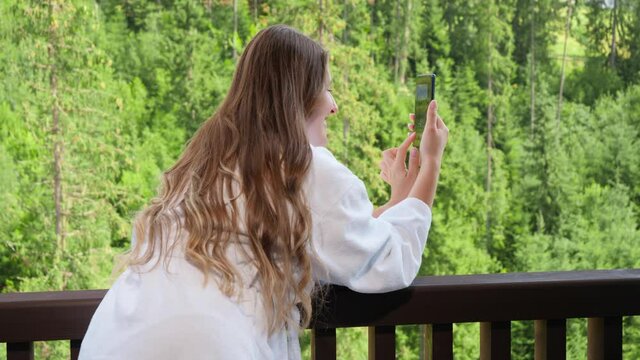 Beautiful young woman in bathrobe standing on balcony and making image of mountains and forest on her smartphone.