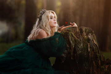 A beautiful blonde young woman in a long green dress and a diadem on her head in the forest. girl sitting near the old stump with butterfly on her hand. Solar glare. Fantasy. fairy tale © Andriy Medvediuk
