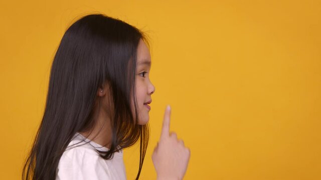 Profile portrait of little asian girl showing quiet gesture aside at free space, orange studio background
