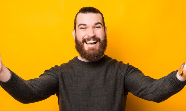 Joyful bearded man taking selfie with camera over yellow background.