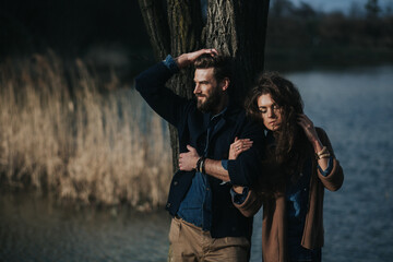Fototapeta na wymiar two caucasian lovers are standing near tree by the lake. Young couple is hugging on autumn day outdoors. A bearded man and curly woman in love. Valentine's Day. Concept of love and family.