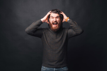 Photo of angry bearded man standing over dark background holding head and screaming.