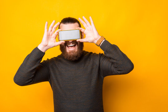 Photo of joyful bearded hipster man holding smartphone over eyes.