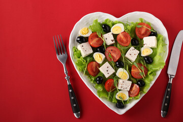 Healthy salad with cherry tomatoes, feta, quail eggs, black olives and microgreens peas on a heart-shaped plate on a red background, Top view
