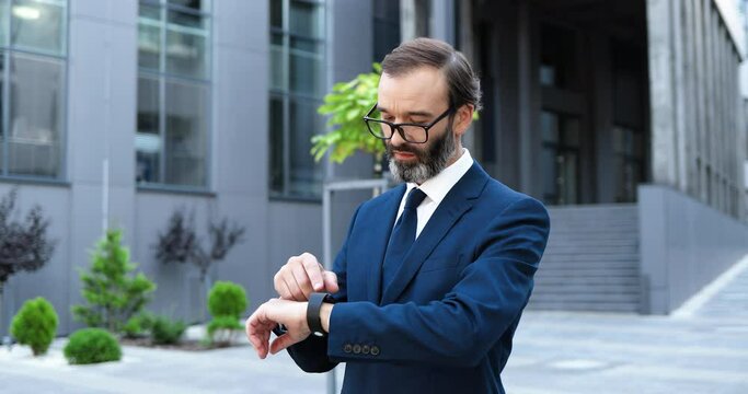 Handsome elegant Caucasian businessman standing outdoor at street and tapping smart watch. Middle-aged man in suit, tie and glasses using device on hand and scrolling in city.