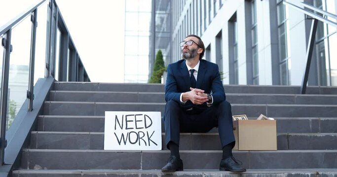 Sad Caucasian Middle-aged Man In Glasses And Tie Sitting On Steps Outdoor. Need Work. Looking For Job. Fired Retired Senior Upset Male. Businessman Loosing Business And Searching Workplace In Crisis.