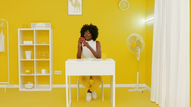 Portrait of stylish African woman sitting at desk in creative yellow office, designed in yellow and white colors, catching red apple that is thrown in the shot, concept of creative video, Slow motion.