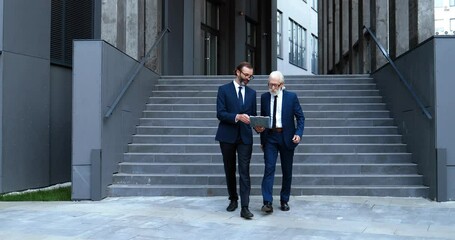 Caucasian man, personal assistant of old businessman walking down the stairs from business center outdoor and showing documents and contract to boss. Males business partners discussing work on walk.