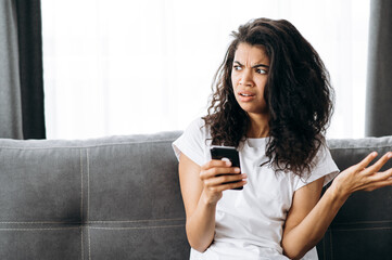 Worried african american girl holding smartphone in hand, got a strange message or news. Confused girl sitting on the couch, browsing internet