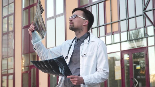 Doctor wearing lab coat stethoscope analyzing X-ray image outdoors stand near hospital main entrance. Young healthcare professional, internet radiologist exam mri x-ray lungs during COVID 19 epidemic