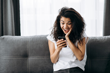 Portrait of beautiful african american young woman at home. The girl sits on the couch and use her phone. Joyful woman look at the screen, reading good news, makes a good purchase online or get a