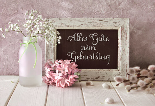 Springtime background. Spring flowers and a blackboard on white table. Pink hyacinth, baby breath and pussy willow. Text Alles gute zum Geburtstag means Happy Birthday.